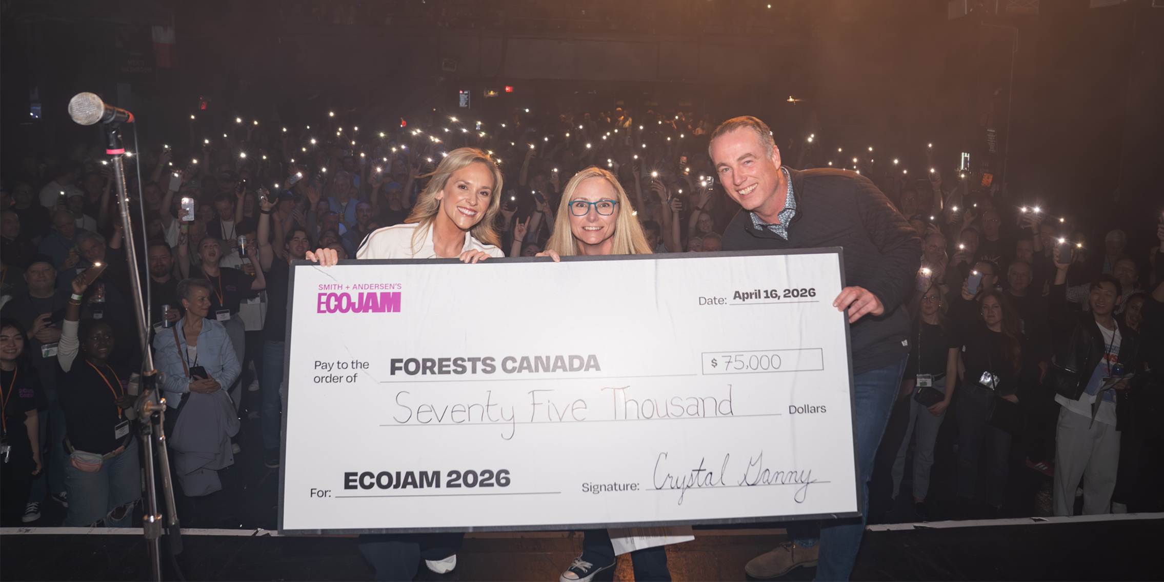 Three people on stage hold a large EcoJam 2026 cheque for $75,000 made out to Forests Canada, with a crowd holding up phone lights in the background.