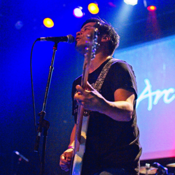 Bassist playing on stage and singing into a mic with blue lighting in the background. 