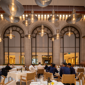 Dining room with patrons, three large arched windows on a heritage facade wall and decorative lighting throughout.
