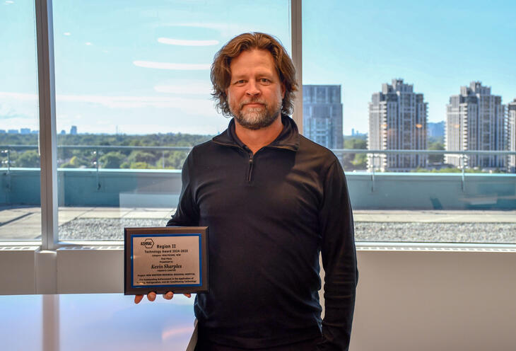 Man in front of a window holding a plaque for the 2024-2025 ASHRAE Technology Awards.