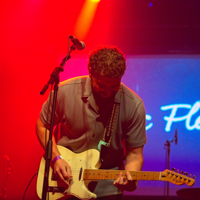 A man with a beard plays an electric guitar on stage under vibrant red and yellow lights. The background features cursive text.