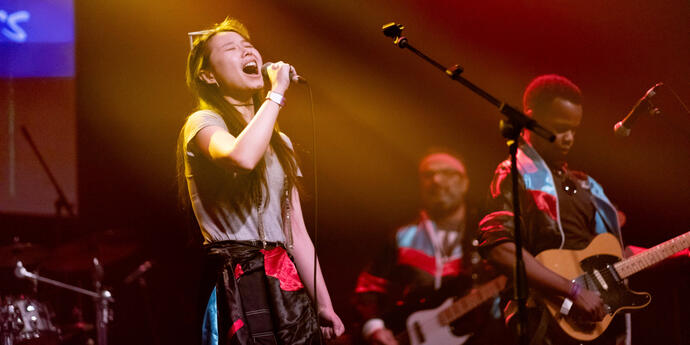 A woman passionately singing into the mic in a yellow spotlight, next to a man looking down at his guitar as he plays.